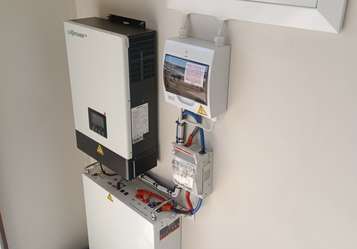 Falra szerelt hibrid inverter, alatta álló lítiumos akkumulátor, mellettük biztosítéktáblák egy lakóépület belső falán